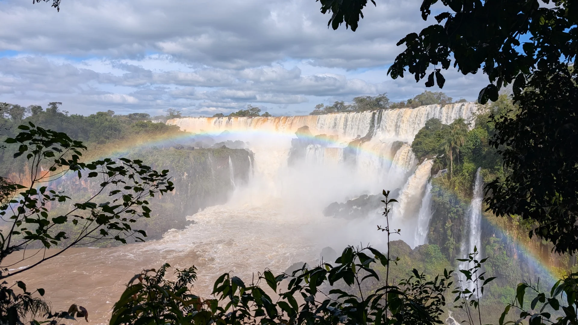 Vista d'un tram ample de les Cascades de l'Iguaçú descarregant aigua amb molta força i creant un espès núvol de boirina. Un arc de Sant Martí molt definit travessa l'escena d'un extrem a l'altre sobre els salts d'aigua. Tota la imatge està emmarcada per fullatge i branques de la selva en primer pla i penya-segats coberts de vegetació sota un cel parcialment ennuvolat.