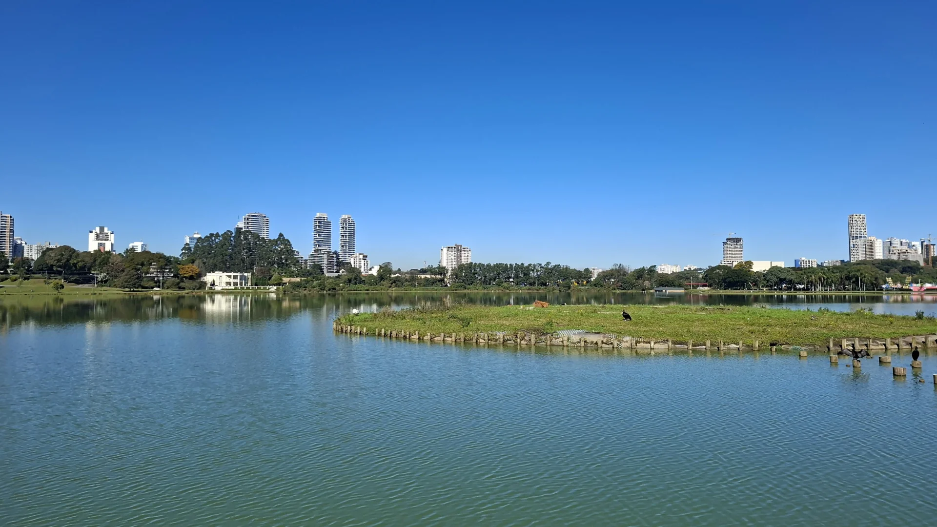 Una vista panoràmica d'un gran llac urbà, amb l'aigua tranquil·la reflectint els edificis de la ciutat al fons sota un cel blau clar, i una illa de vegetació verda al mig del llac.