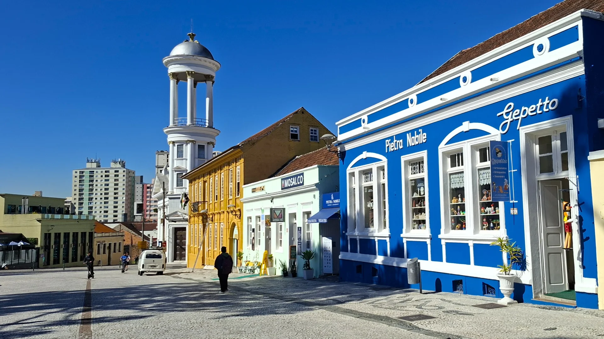 Una vista d'un carrer històric amb edificis colonials de colors variats sota un cel blau, inclòs un edifici blau a la dreta amb un cartell de 'Gepetto' i 'Pietra Nobile', i una estructura de torre blanca al fons.