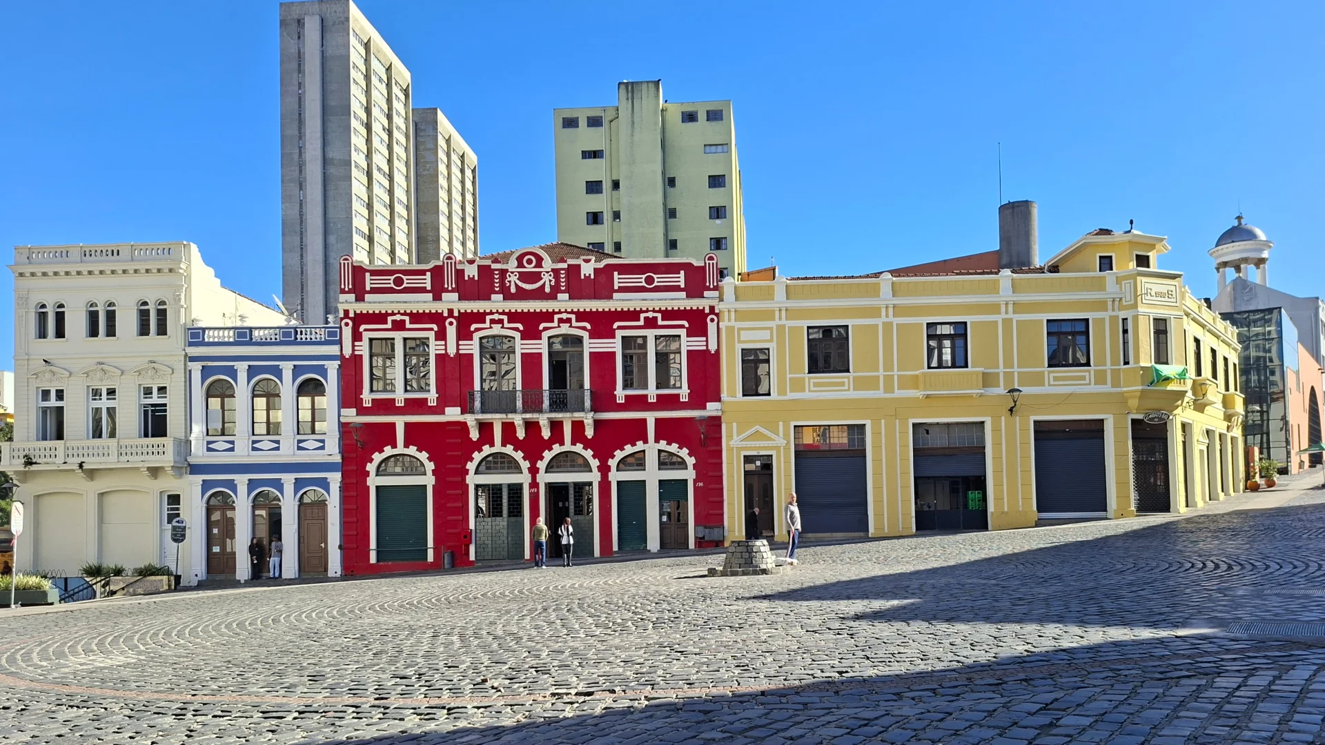 Una vista d'una plaça històrica amb llambordes i una fila d'edificis colonials colorits, inclosos els vermells, grocs i blaus, sota un cel blau clar, amb una petita font de pedra i persones a la plaça.