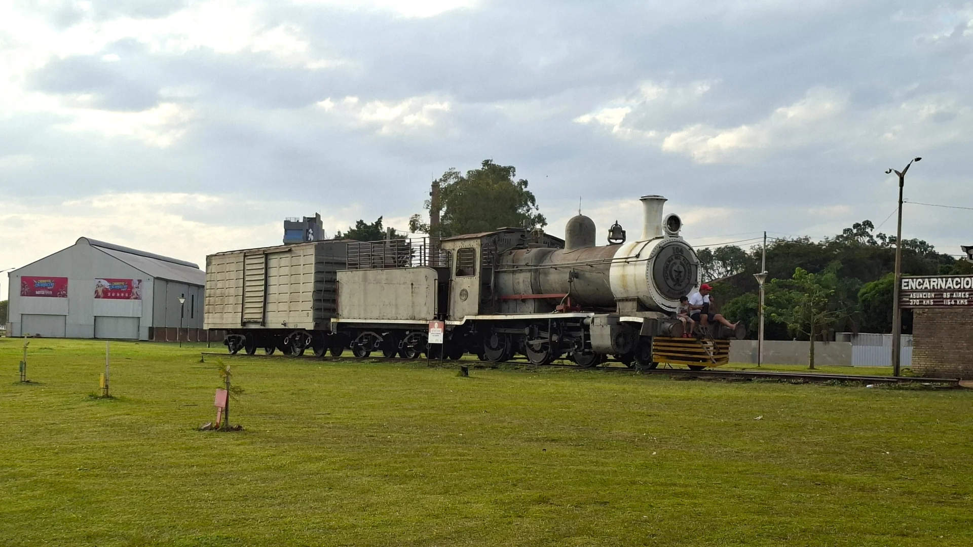 Una antiga locomotora de vapor històrica amb diversos vagons exposada sobre les vies en un ampli prat d'herba verda a Encarnación, amb un parell de persones assegudes a la part frontal de la màquina, un gran cartell indicador de fusta a la dreta i una nau industrial de fons sota un cel ennuvolat.