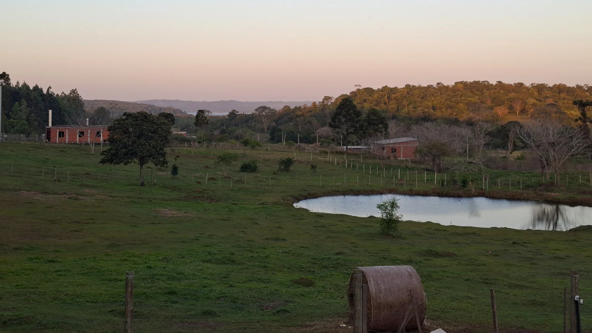 Vista panoràmica de la propietat rural de Hohenau, amb un paisatge de camps verds i turons recoberts de bosc frondós al fons sota un cel crepuscular amb tons suaus. En primer pla, una tanca de fusta i un objecte cilíndric de palla sobre el prat, amb un estany d'aigua tranquil·la a la dreta i construccions rústiques a l'esquerra.