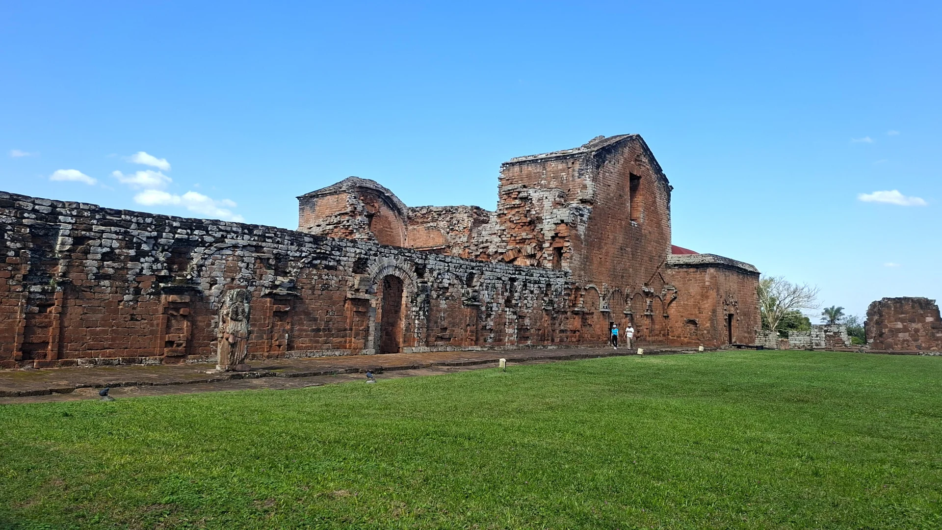Vista panoràmica dels majestuosos murs de pedra de l'antiga església a la reducció jesuïta de Santíssima Trinitat del Paraná, amb arcs i estructures adossades a la paret de pedra vermella, estenent-se al costat d'un ampli prat d'herba verda sota un cel blau amb núvols suaus i dues persones passejant a la vora.