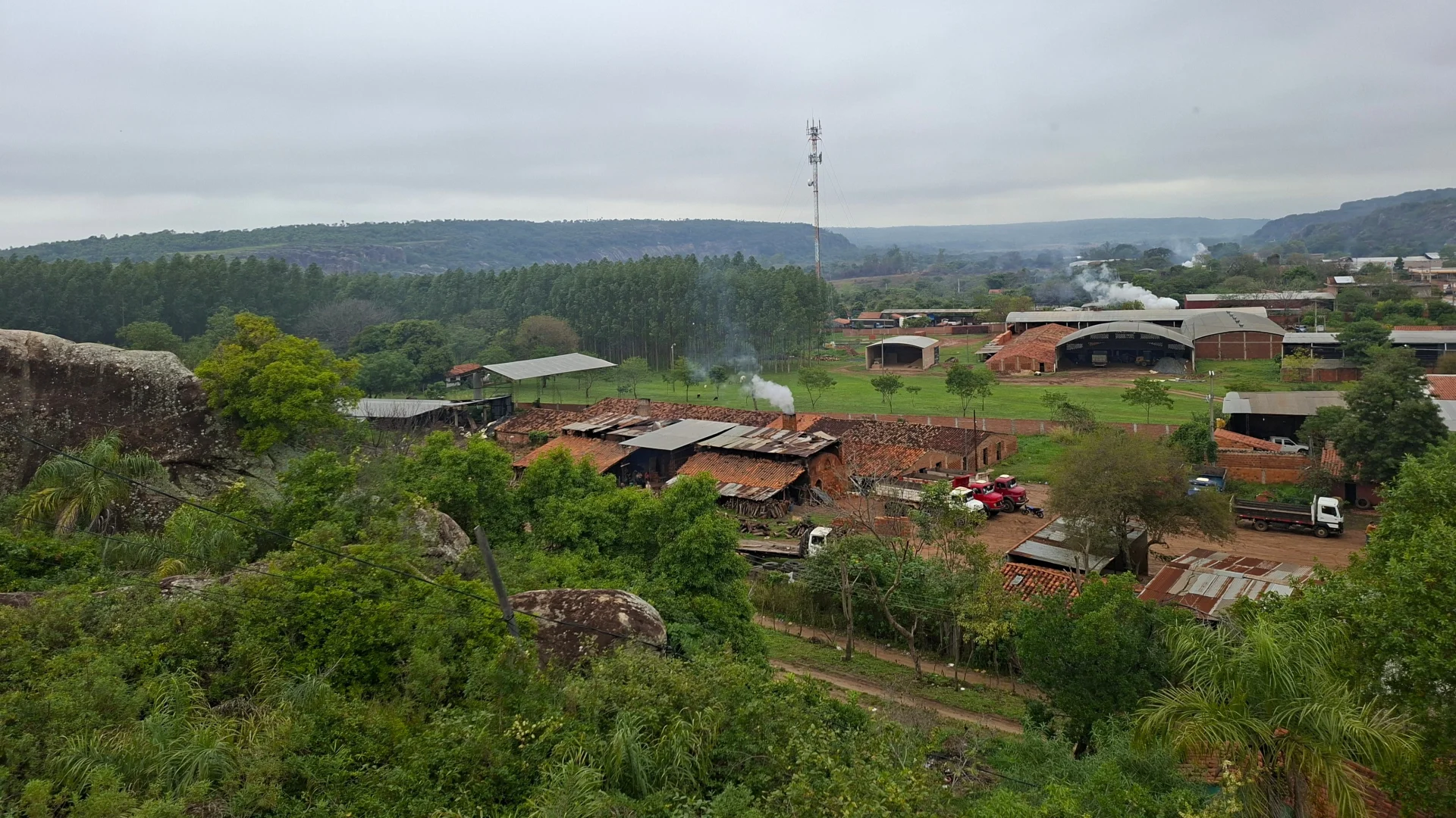 Vista panoràmica elevada del paisatge rural de Tobatí sota un cel ennuvolat, on s'observen diverses indústries tradicionals de ceràmica i maons amb teulades rústiques traient fum, envoltades de camins de terra amb camions estacionats, turons verds i una alta antena de telecomunicacions al fons.