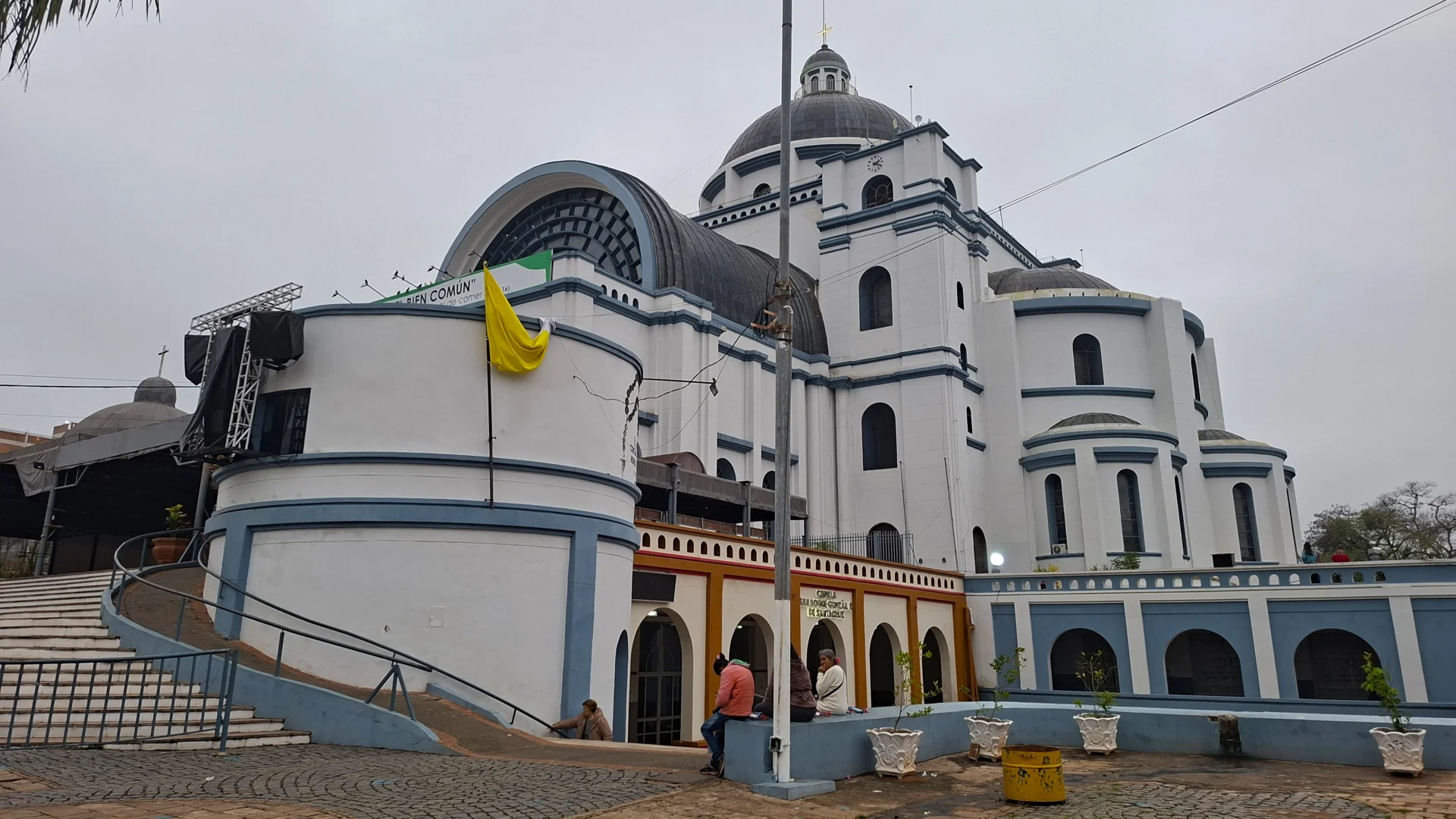 Vista exterior de la monumental Basílica de la Mare de Déu dels Miracles a Caacupé, un temple d'estil singular amb façanes pintades de blanc-i-blau, grans cúpules i cossos arrodonits, amb una bandera groga i blanca penjant de la barana, escalinates laterals i algunes persones descansant a la plaça inferior sota un cel encotxat.
