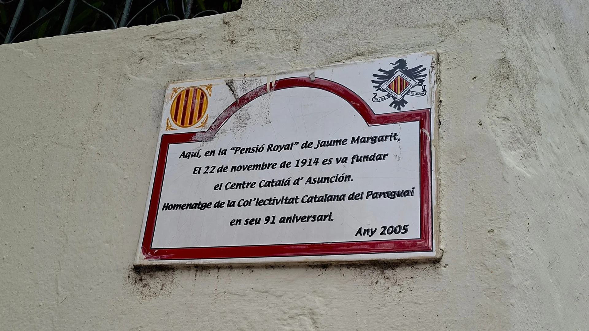 Primer pla d'una placa commemorativa de ceràmica sobre una paret clara a Asunción, encapçalada per la senyera catalana i el logotip de l'entitat, amb una inscripció que recorda la fundació del Centre Català d'Asunción el 22 de novembre de 1914 a la Pensió Royal de Jaume Margarit.
