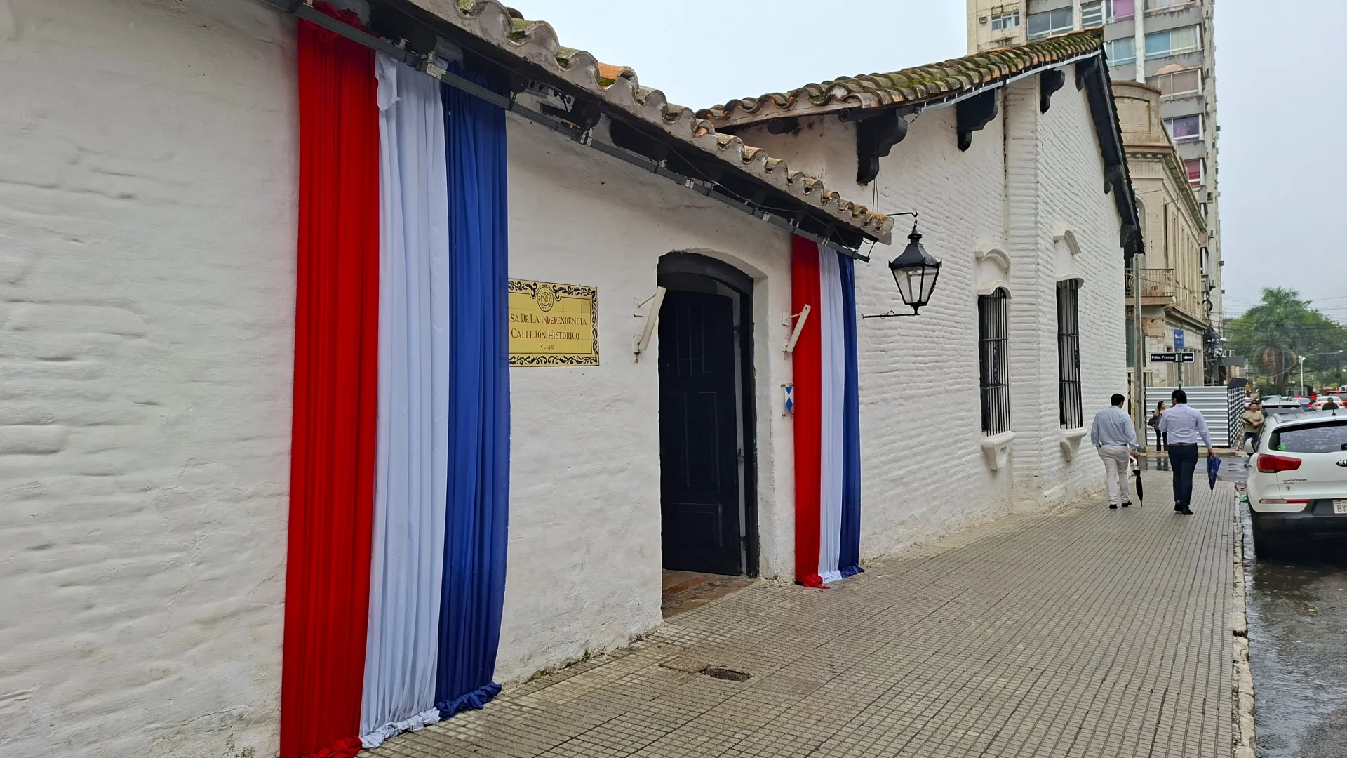 Façana d'estil colonial de la Casa de la Independència a Asunción, amb parets blanques, teulada tradicional i dues grans teles verticals amb els colors vermell, blanc-i-blau de la bandera del Paraguai emmarcant la porta principal de fusta, acompanyada d'una placa daurada i un fanal de ferro forjat a la vorera.