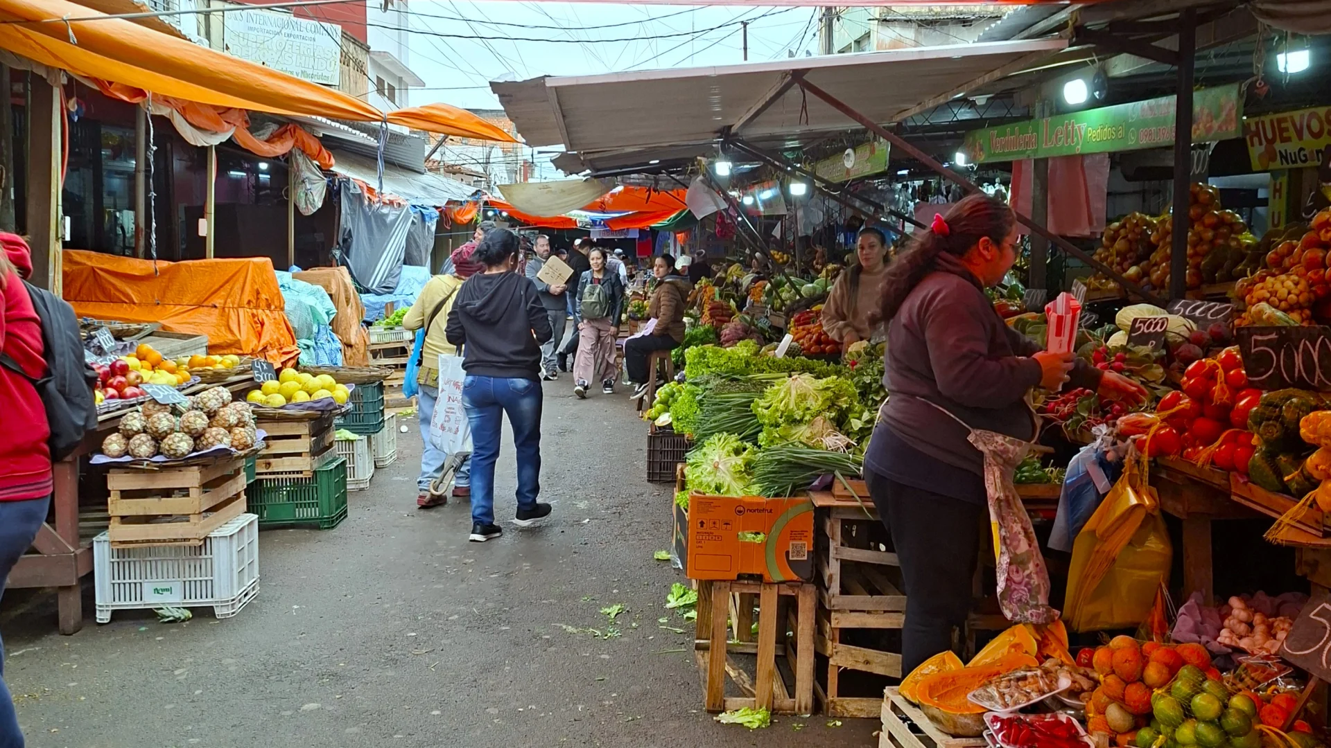 Vista d'un carreró molt concorregut del Mercado 4 a Asunción, amb tendals ataronjats i blancs cobrint nombroses paradetes a l'aire lliure, caixes de fusta plenes de fruita i verdura fresca com pinyes, taronges, carabasses i verdures, amb compradors caminant entre el passadís.