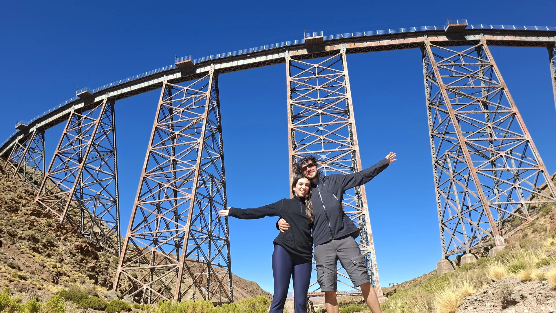 La Irene i en Pau somrients amb els braços oberts sota l'espectacular viaducte de La Polvorilla a San Antonio de los Cobres. L'alta estructura metàl·lica d'acer amb pilars d'armadura s'alça cap al cel blau intens per sobre del terreny àrid de la puna.