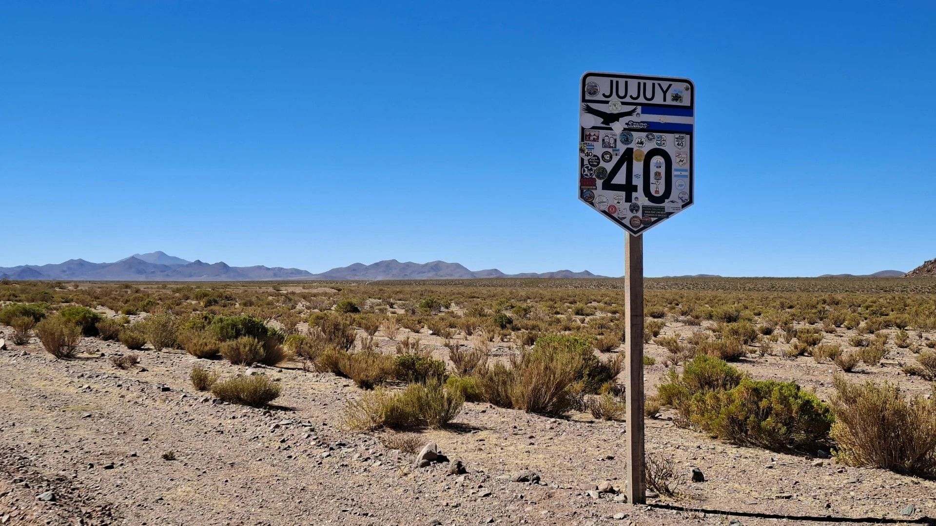 Primer pla d'un cartell vial metàl·lic de la Ruta 40 ple d'adhesius de viatgers al seu pas per Jujuy, clavat sobre un pal de fusta al voral d'una pista de terra. Al fons s'estén la immensitat de la plana desèrtica amb petits matolls i muntanyes a l'horitzó sota un cel blau lluent.