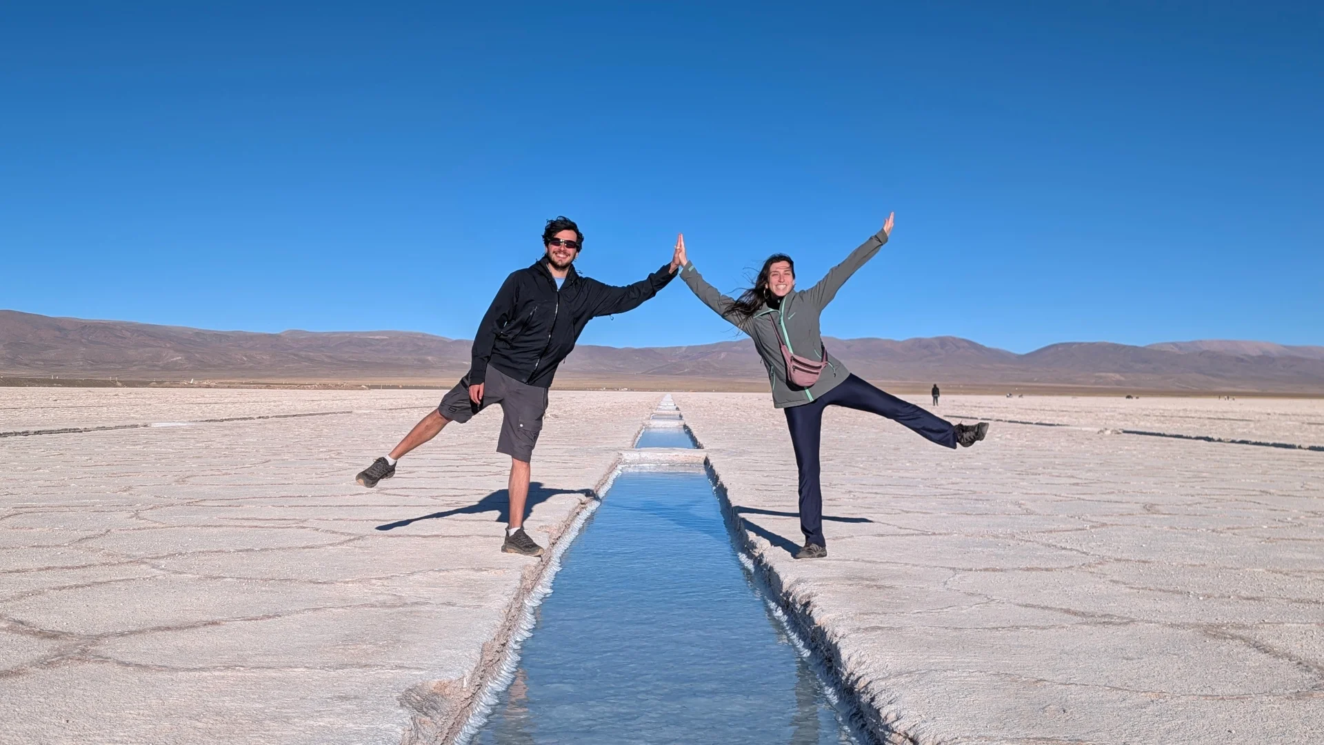 En Pau i la Irene fent una divertida postura d'equilibri sobre la crosta de sal blanca de les Salinas Grandes, tocant-se les mans amb un