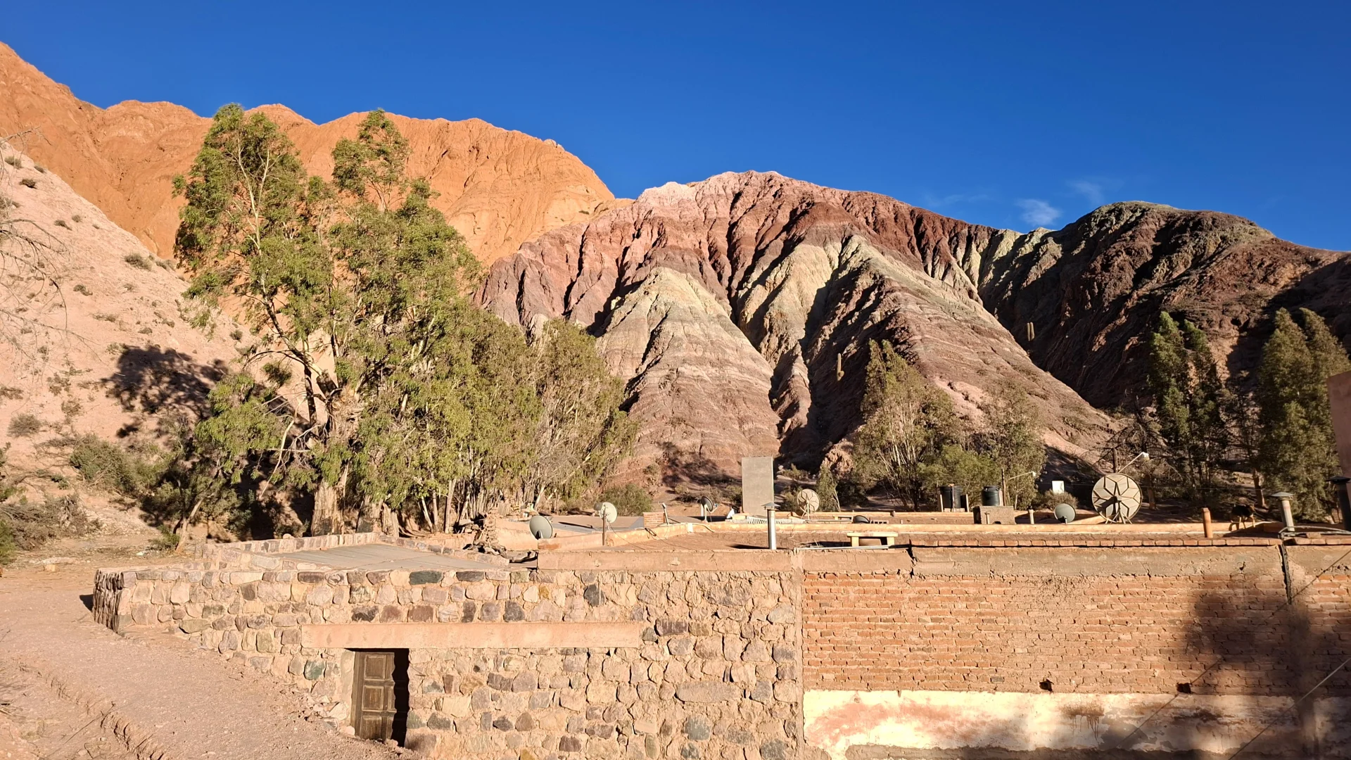 Vista del Cerro de los Siete Colores a Purmamarca, amb els seus estrats de roca sedimentària de colors vermellosos, grocs, verds i violacis alçant-se darrere de teulades i cases tradicionals d'adob i maó, amb arbres verds sota un cel blau intens.