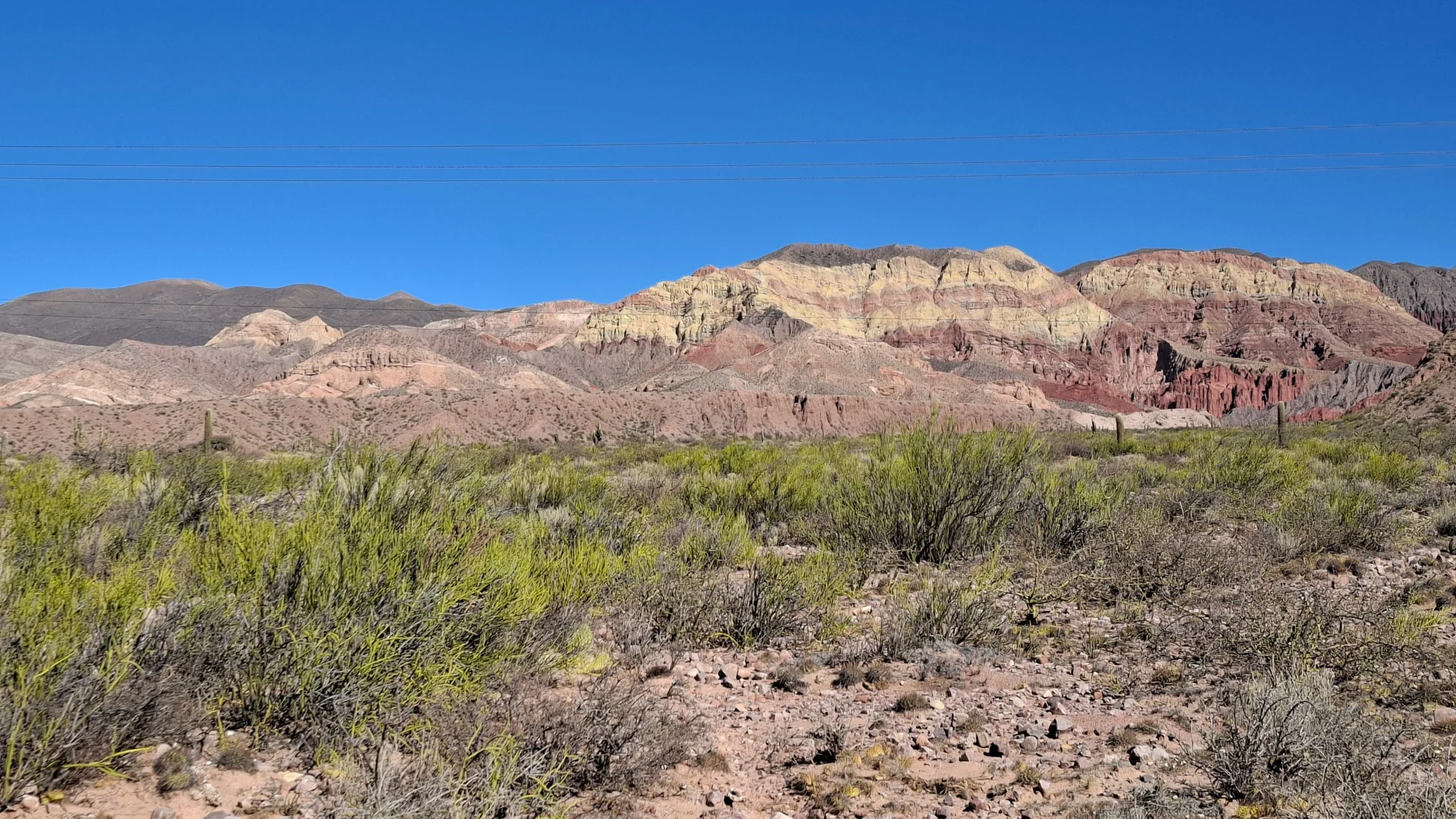 Vista panoràmica del paisatge àrid de la Quebrada de Humahuaca en direcció a Uquía, amb una plana de matolls i vegetació baixa en primer pla i una impressionant serralada al fons amb capes estratificades de colors groc, vermell i ocre sota un cel blau net.