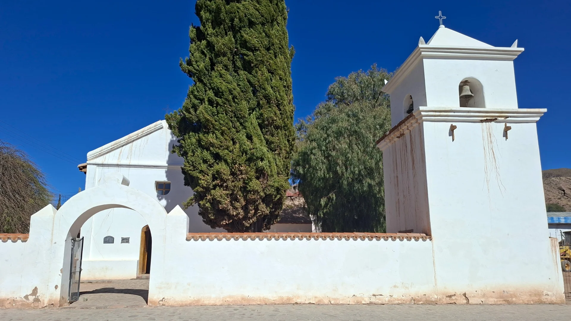 Façana exterior de la històrica església de San Francisco de Paula a Uquía, pintada d'un blanc immaculat, amb la seva torre campanar exempta amb campanes de bronze, un mur perimetral amb arc d'entrada de mig punt i un alt xiprer verd al pati sota un cel blau radiant.