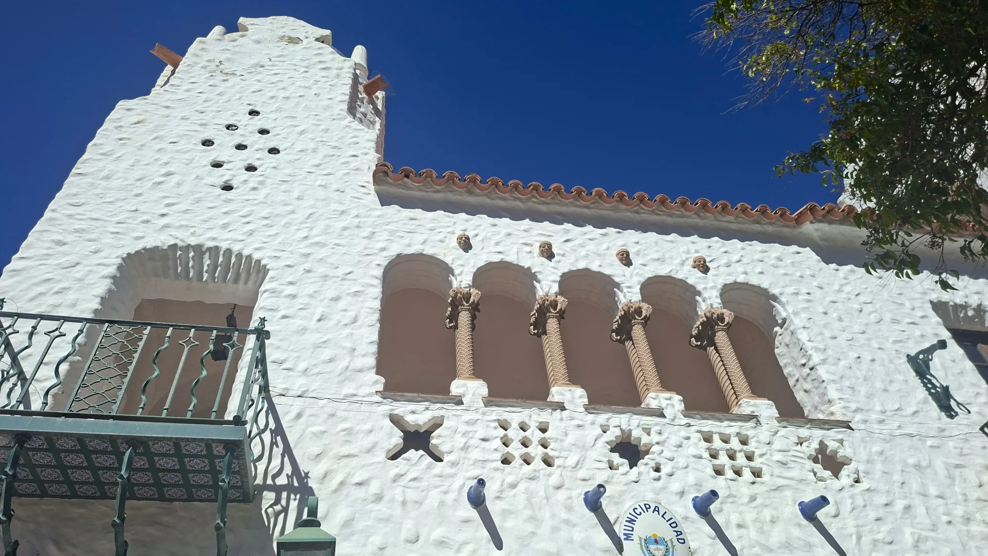 Detall arquitectònic de la façana emblanquinada del Cabildo de Humahuaca, mostrant una galeria d'arcs amb columnes salomòniques decorades, petits rostres esculpits en relleu a la paret, un balcó de ferro forjat a l'esquerra i l'escut municipal a la part inferior sota un cel blau.