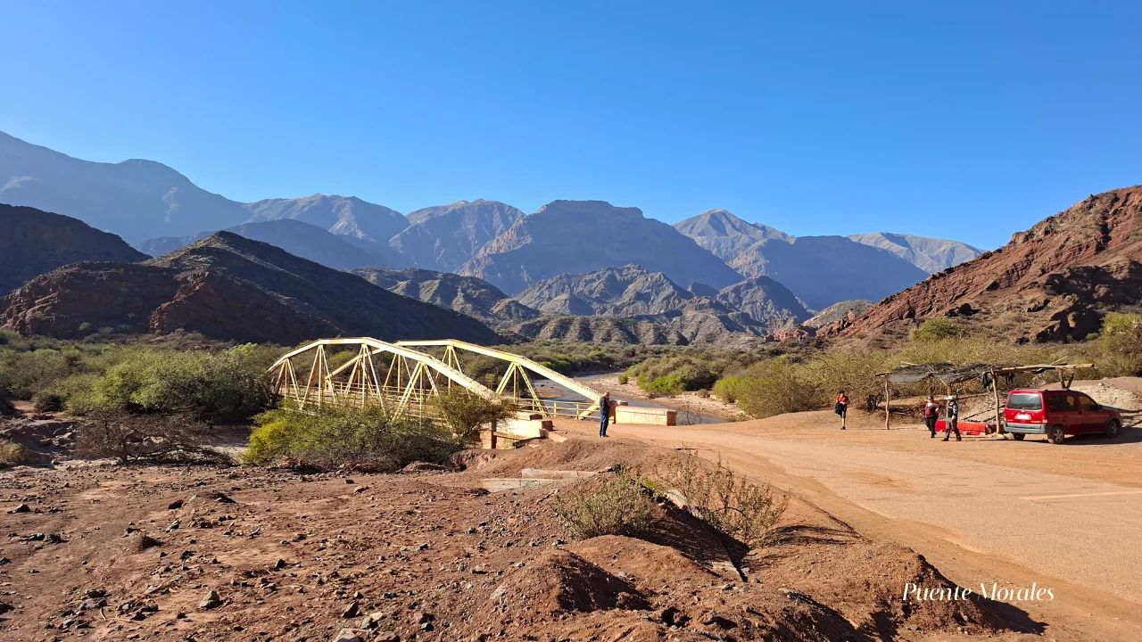 Vista panoràmica del Puente Morales a la Quebrada de las Conchas, amb un pont metàl·lic de color groc que creua la llera seca d'un riu sobre un camí de terra vermellosa, flanquejat per espectaculars serralades de roca vermella i muntanyes escarpades sota un cel blau intens, amb una furgoneta vermella i visitants a la dreta.