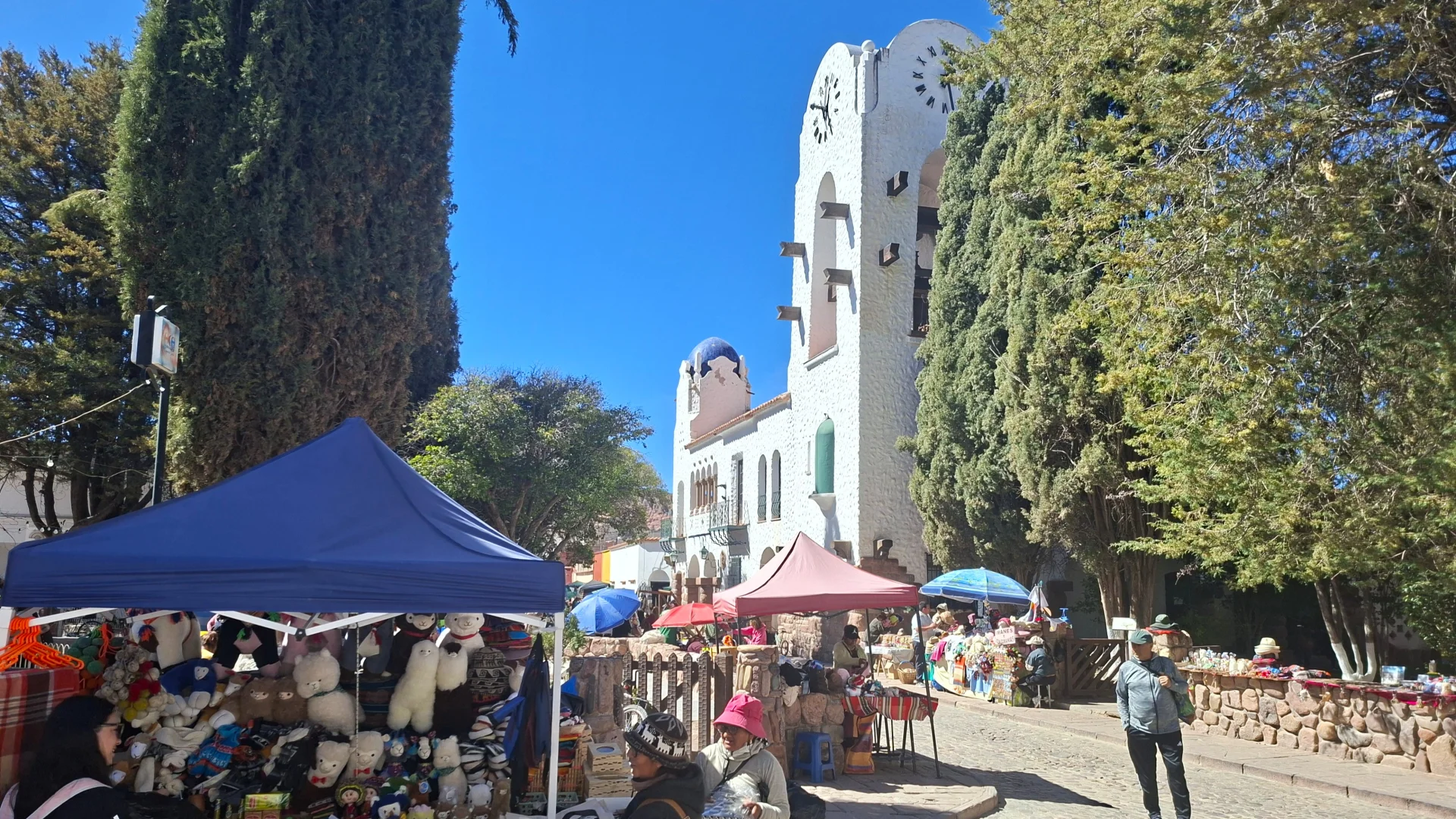 Vista de la plaça d'Humahuaca davant del Cabildo en un dia assolellat, amb parades d'artesania sota tendals blaus i vermells plenes de ninots de llana de llama i teixits tradicionals, amb gent passejant i la torre blanca del rellotge de l'ajuntament alçant-se entre grans xiprers.
