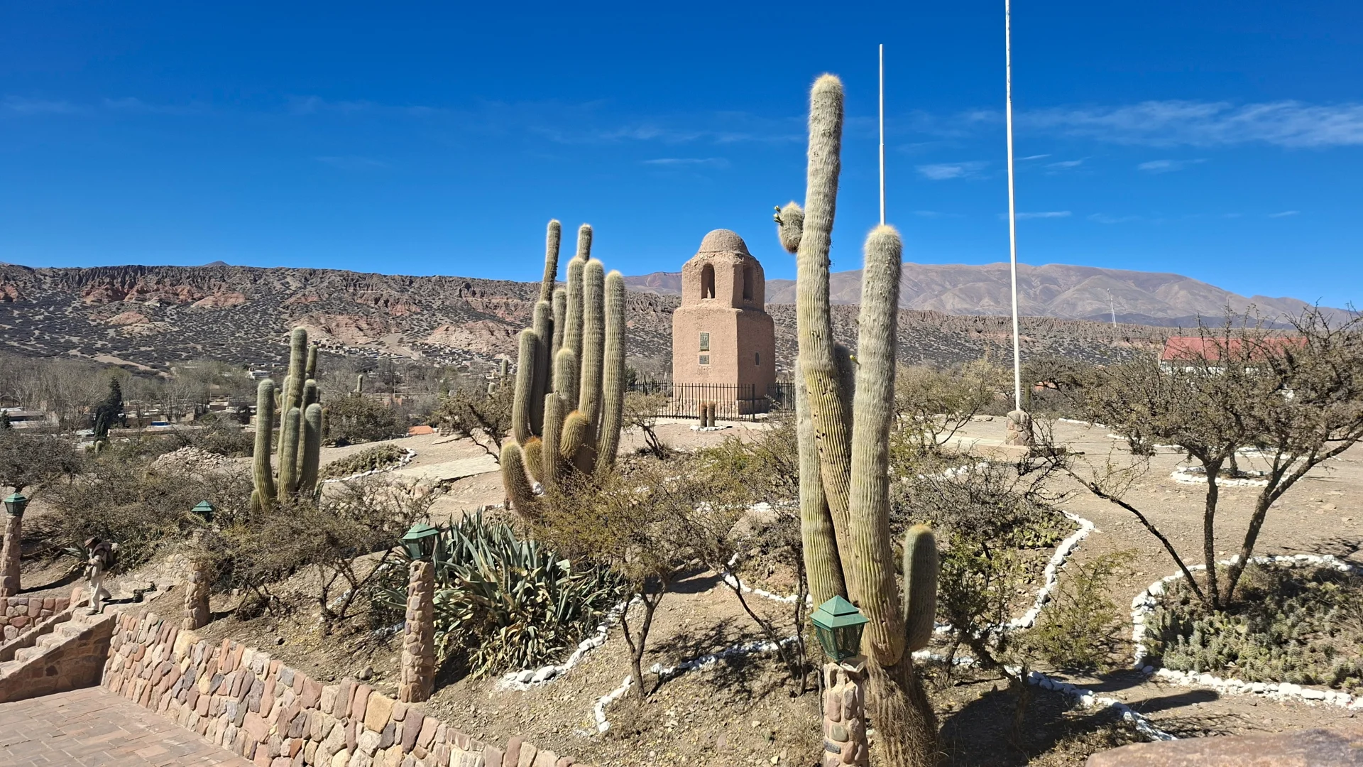 Vista de la històrica Torre de Santa Bàrbara a Humahuaca, una edificació d'adob amb cúpula sobre un turó àrid envoltat de cactus cardons gegants, atzavares i fanals de pedra, amb una cadena muntanyosa al fons sota un cel blau net.