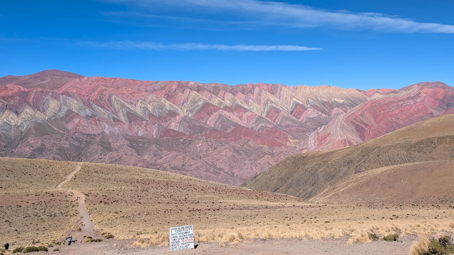 Vista panoràmica de la Serranía del Hornocal (la muntanya dels 14 colors) a Humahuaca, amb els seus plecs geològics triangulars en forma de ziga-zaga en tons vermells, grocs i ocres, un sender que descendeix per la puna i un cartell informatiu en primer pla sota un cel blau clar.