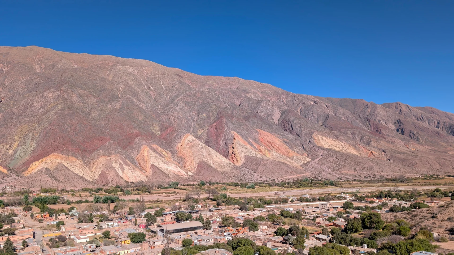 Vista panoràmica en alçada del poble de Maimará a la Quebrada de Humahuaca, amb les cases de la localitat en primer pla i la formació de la Paleta del Pintor de fons, mostrant les seves franges ondulades de colors grocs, ataronjats i vermellosos sota un cel blau net.