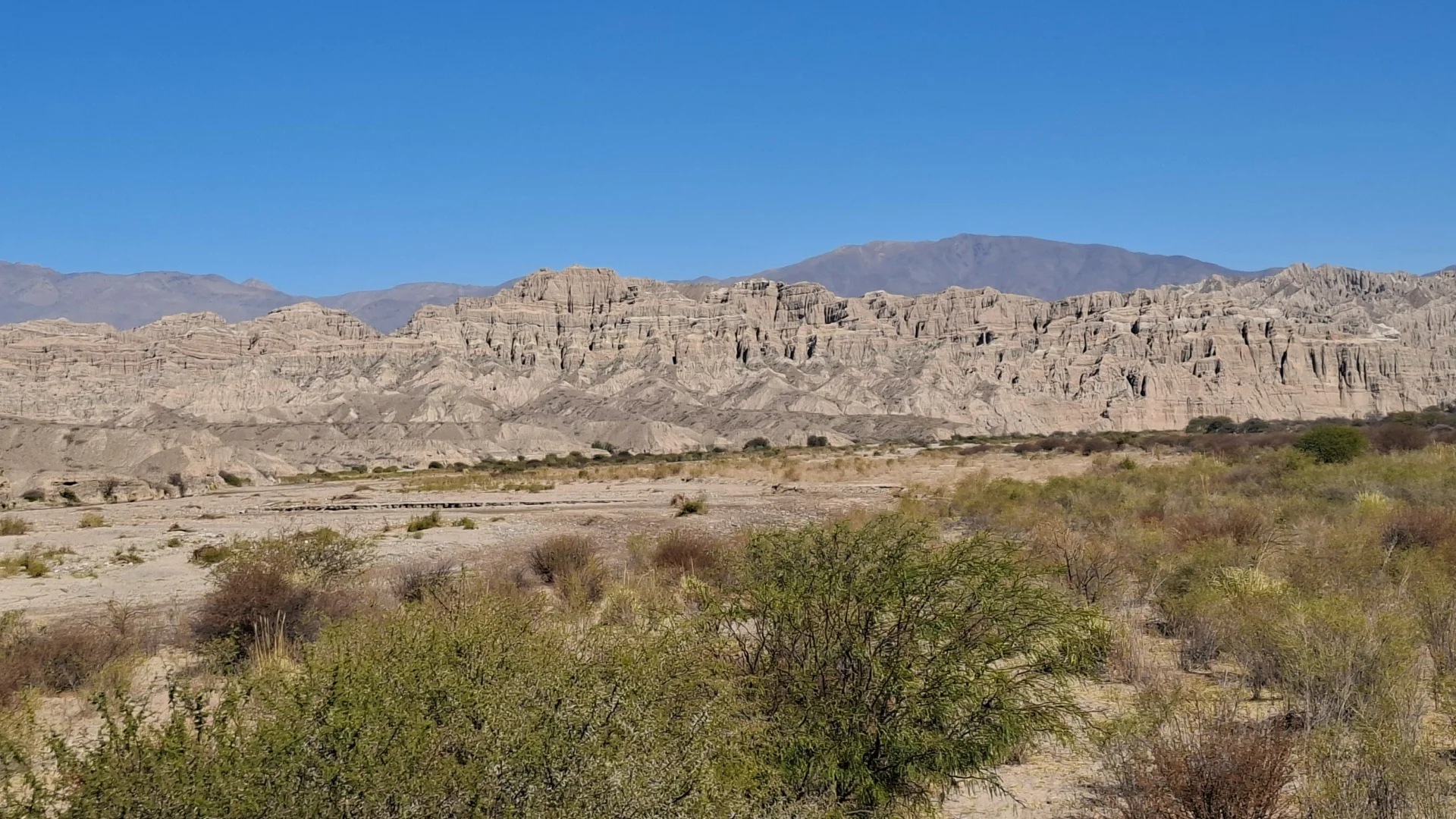 Vista panoràmica dels paisatges erosionats de la Quebrada de las Flechas, amb una extensa plana de matolls i vegetació baixa en primer pla i una singular formació de crestes rocoses punxegudes i inclinades que s'alcen davant d'una muntanya llunyana sota un cel blau immaculat.