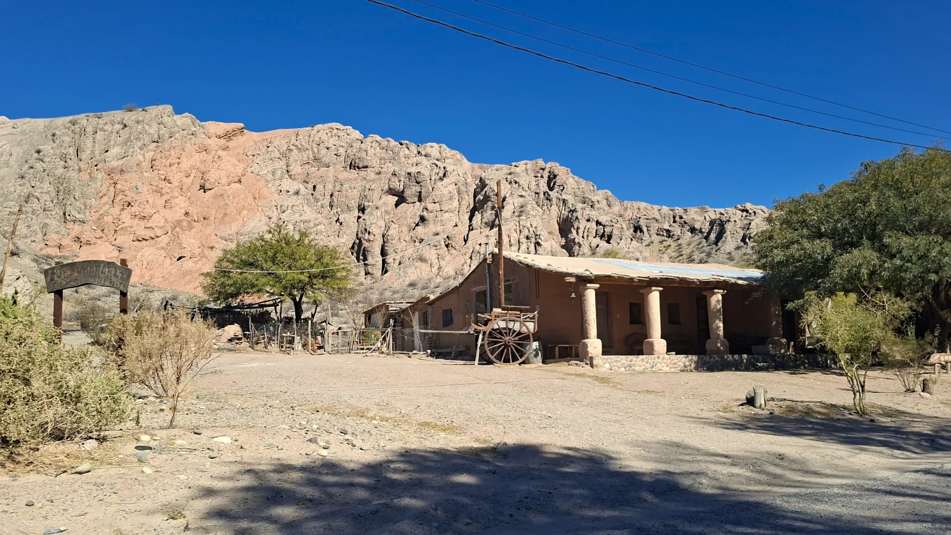 Vista d'una casa tradicional d'adob amb porxo sostingut per columnes rústiques i un carro de fusta amb grans rodes estacionat al davant, situada al costat d'un camí de terra amb una imponent serralada de roca vermellosa alçada just al darrere sota un cel blau net.
