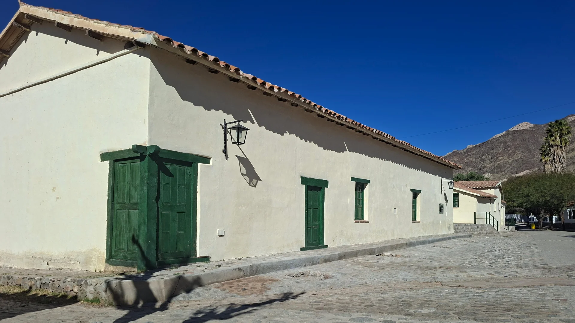 Vista exterior d'una clàssica casa colonial a Seclantás, amb façana emblanquinada, teulada de teules vermelles, finestres i portes de fusta pintades de verd intens, fanals d'època i un carrer empedrat sota un cel blau profund amb una muntanya àrida al fons.