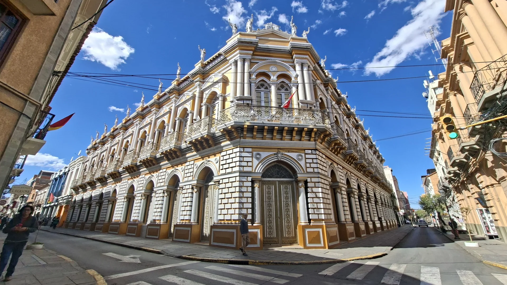 Vista exterior en perspectiva de la imponent façana de la Casa Dorada en una cantonada de Tarija sota un cel blau amb núvols dispersos. La mansió senyorial llueix detalls d'estil art nouveau en tons ocres i blancs, amb galeries d'arcs a la planta baixa, balcons amb reixes de ferro forjat, elegants columnes i un seguit d'estàtues clàssiques coronant la cornisa superior sobre un carrer empedrat.