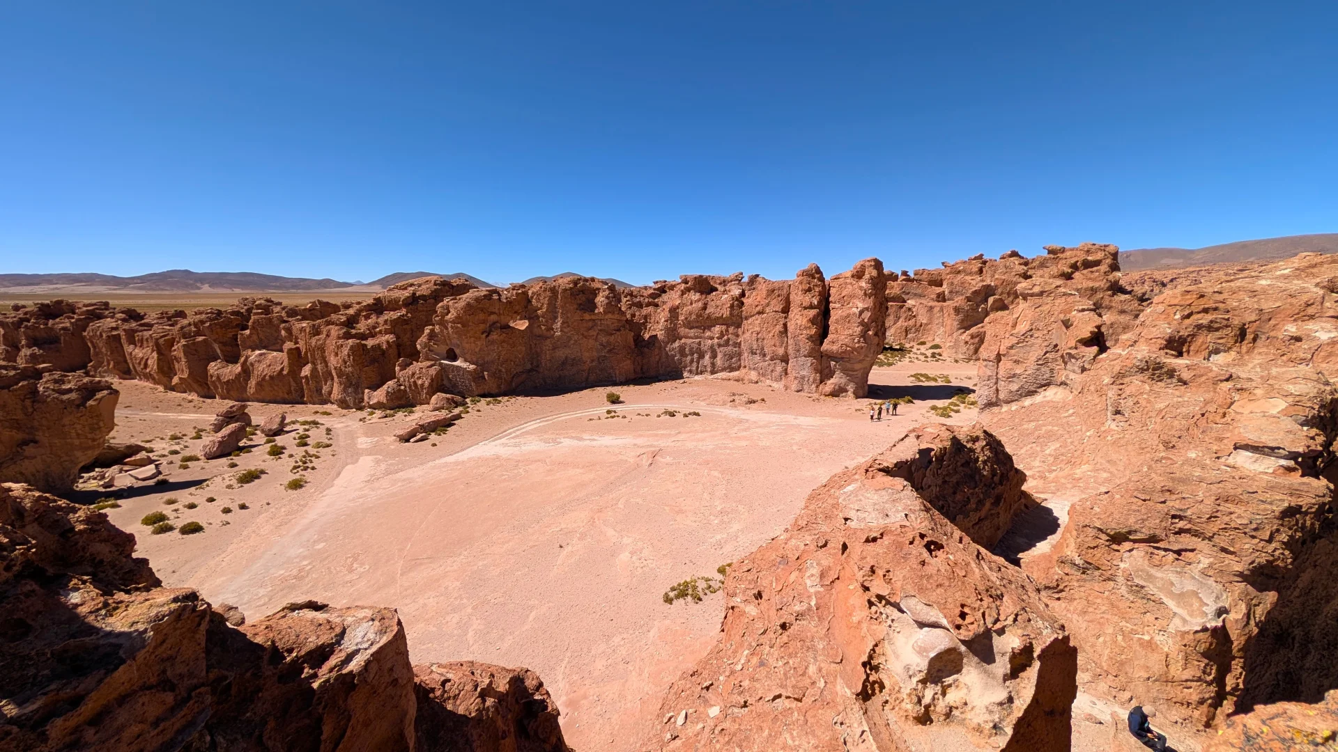 Vista panoràmica del laberint rocós de la Itàlia Perduda, que mostra una gran depressió circular de sorra envoltada per imponents murallons i pinacles de roca volcànica vermellosa, amb petits visitants a la base i muntanyes llunyanes sota un cel blau.