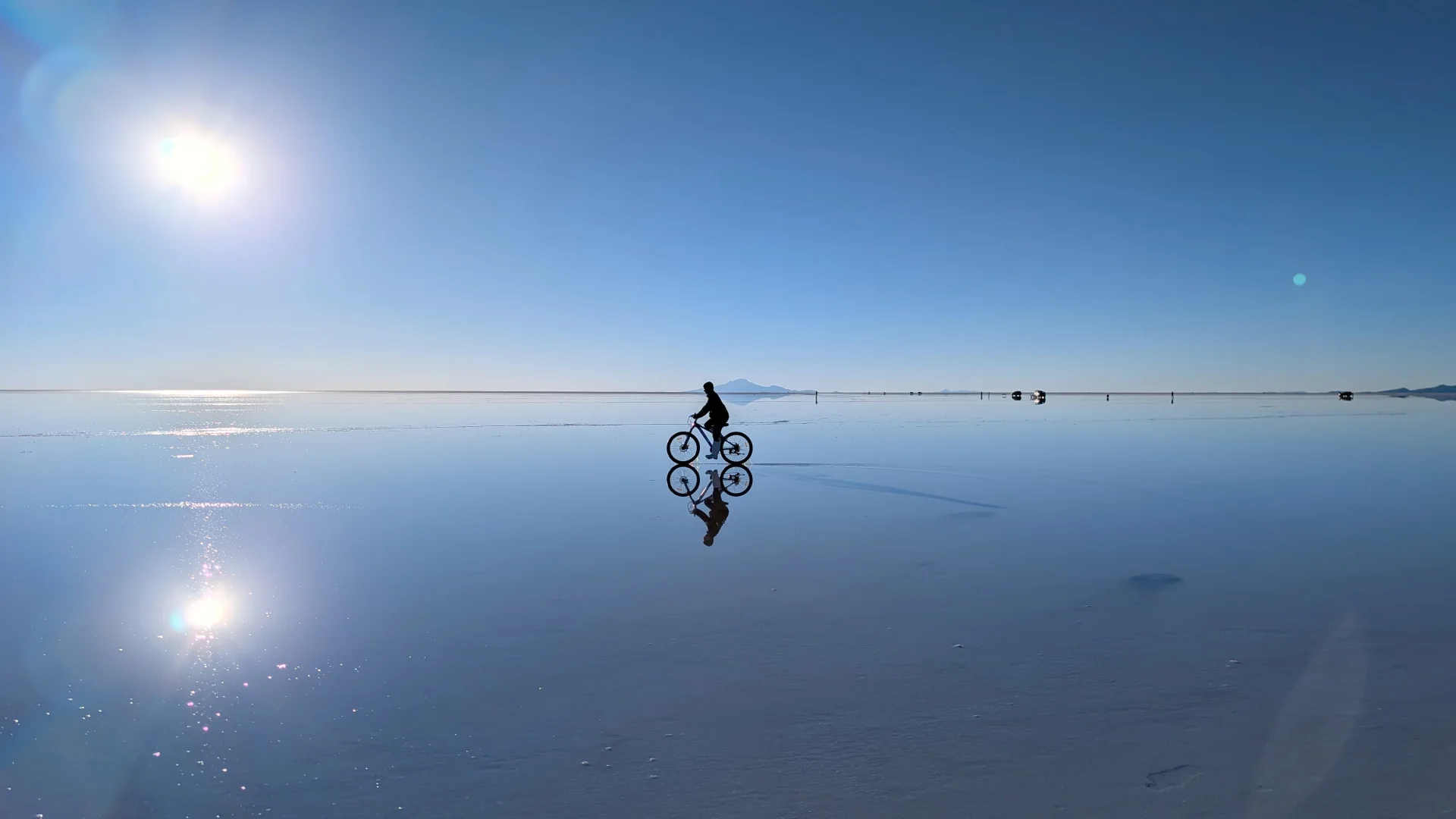 Silueta d'una persona pedalant en bicicleta sobre la superfície inundada del Salar d'Uyuni, creant un perfecte efecte mirall a la làmina d'aigua cristal·lina, sota un sol radiant que produeix un reflex lluent al cel blau.