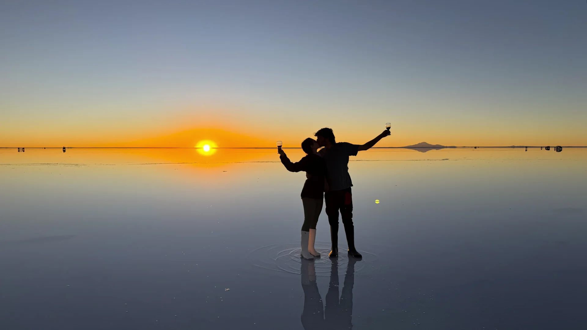 Silueta a contrallum de la Irene i en Pau fent-se un petó i brindant amb copes de vi sobre l'aigua del Salar d'Uyuni durant el capvespre, amb el disc solar amagant-se a l'horitzó i tenyint el cel i el reflex de l'aigua de càlids tons ataronjats i daurats.