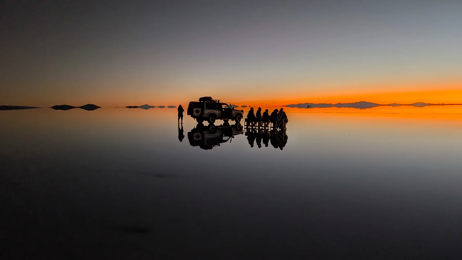 Vista panoràmica en fer-se fosc al Salar d'Uyuni, amb les siluetes del vehicle 4x4 i el grup de viatgers asseguts i drets reflectides nítidament sobre la làmina d'aigua infinita, retallades contra un cel crepuscular amb un degradat de taronja intens a blau fosc.