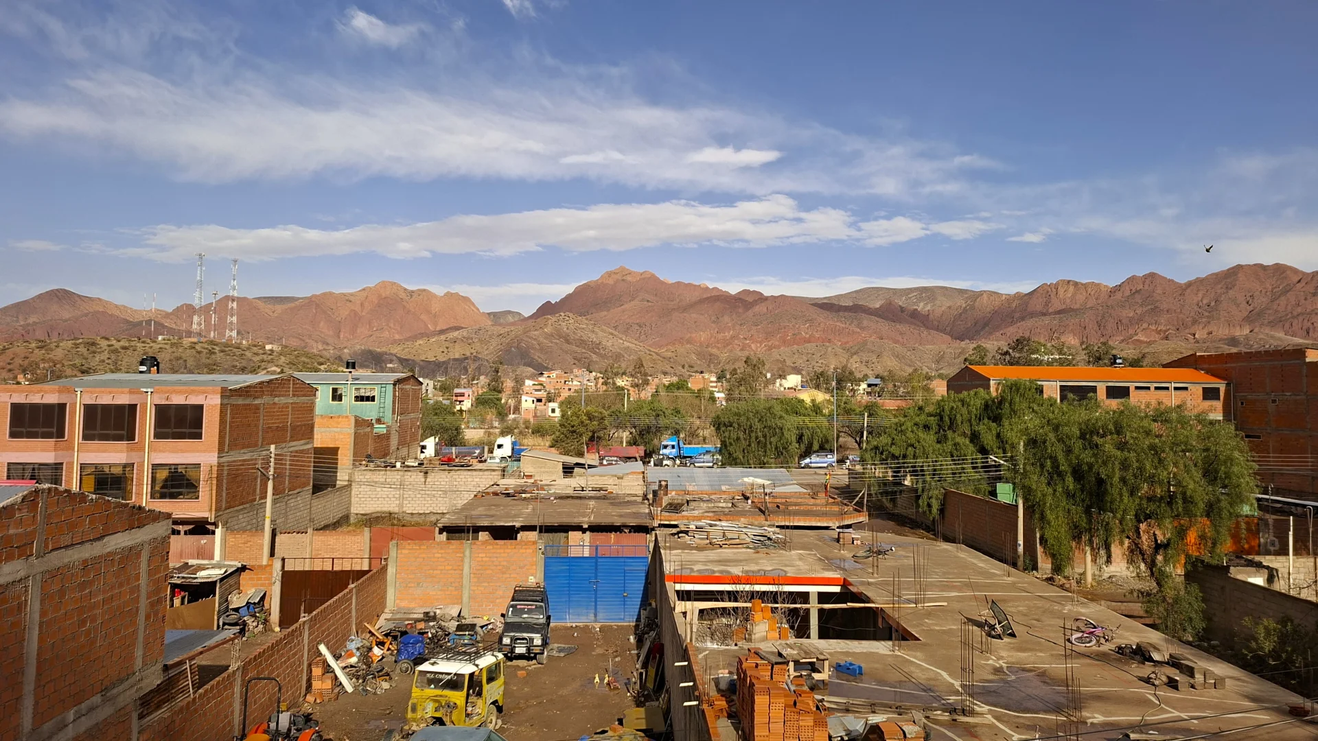Vista panoràmica elevada des de Tupiza sobre els terrats i patis de construcció de maó del poble, amb una imponent serralada de muntanyes i turons vermellosos i escarpats alçant-se al fons sota un cel blau amb núvols suaus.  setmana30_foto2: Tram d'una carretera de muntanya asfaltada serpentejant a través d'un pas d'altura cap a Uyuni, amb clapes de neu i gebre cobrint la terra i els matolls del voral i turons ondulats enfarinats de neu al fons sota un cel clar.