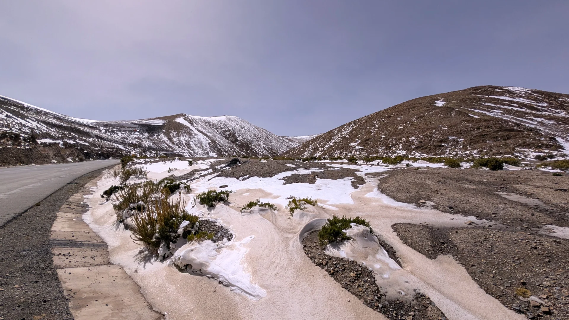 Tram d'una carretera de muntanya asfaltada serpentejant a través d'un pas d'altura cap a Uyuni, amb clapes de neu i gebre cobrint la terra i els matolls del voral i turons ondulats enfarinats de neu al fons sota un cel clar.