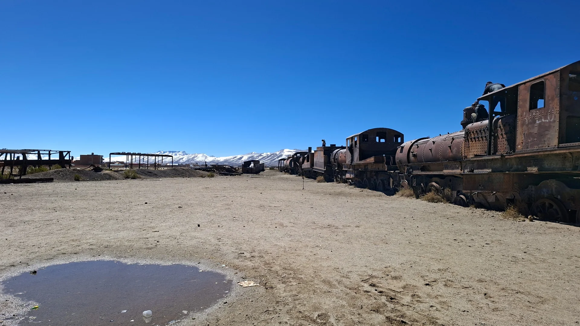 Vista del cementiri de trens d'Uyuni sota un cel blau intens, amb una llarga filera de velles locomotores de vapor i vagons rovellats abandonats sobre la plana sorrenca, un petit bassal d'aigua en primer pla i muntanyes nevades a l'horitzó llunyà.