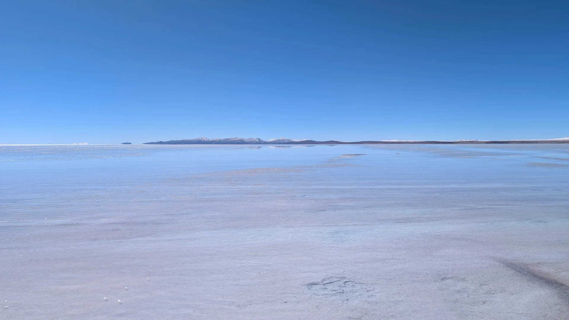 Vista panoràmica de la immensitat inundada del Salar d'Uyuni, on una fina làmina d'aigua sobre la crosta de sal blanca crea un gegantí efecte mirall reflectint el cel blau radiant fins a les siluetes llunyanes de les muntanyes a l'horitzó.