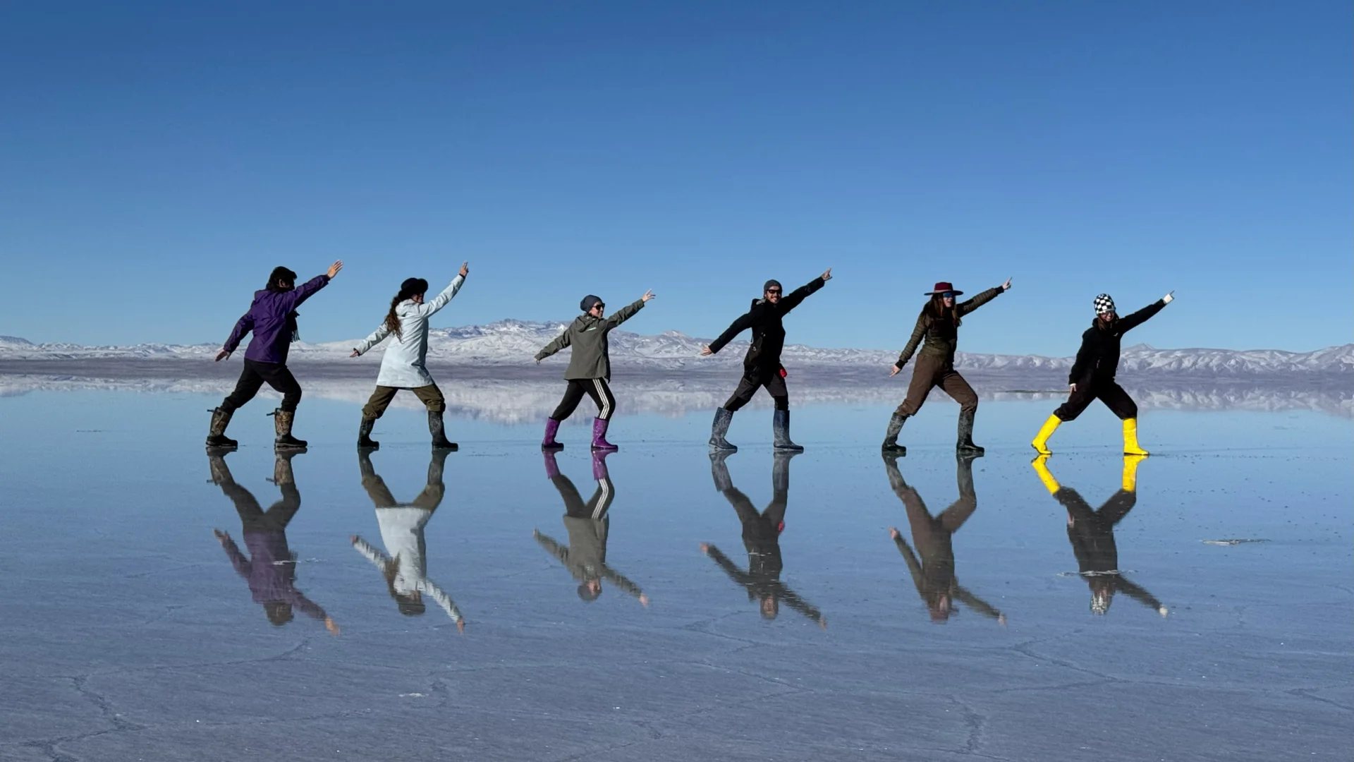 El grup de sis viatgers, incloent-hi la Irene i en Pau, fent una divertida posa coordinada en línia amb un braç alçat sobre la làmina d'aigua del Salar d'Uyuni, amb els seus reflexos nítids projectats sobre l'aigua i muntanyes nevades al fons sota un cel blau net.