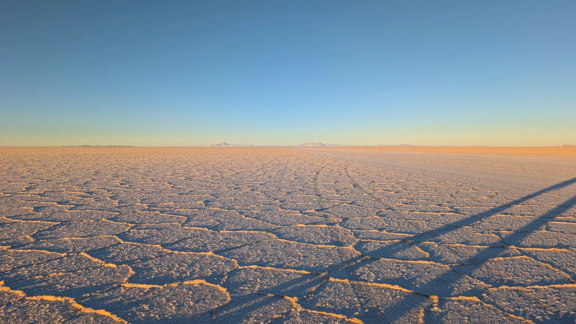 Vista de la superfície seca del Salar d'Uyuni al capvespre, mostrant el característic patró geomètric dels polígons de sal ressaltats per la càlida llum daurada de la posta de sol i llargues ombres sobre la crosta blanca amb les muntanyes a l'horitzó.
