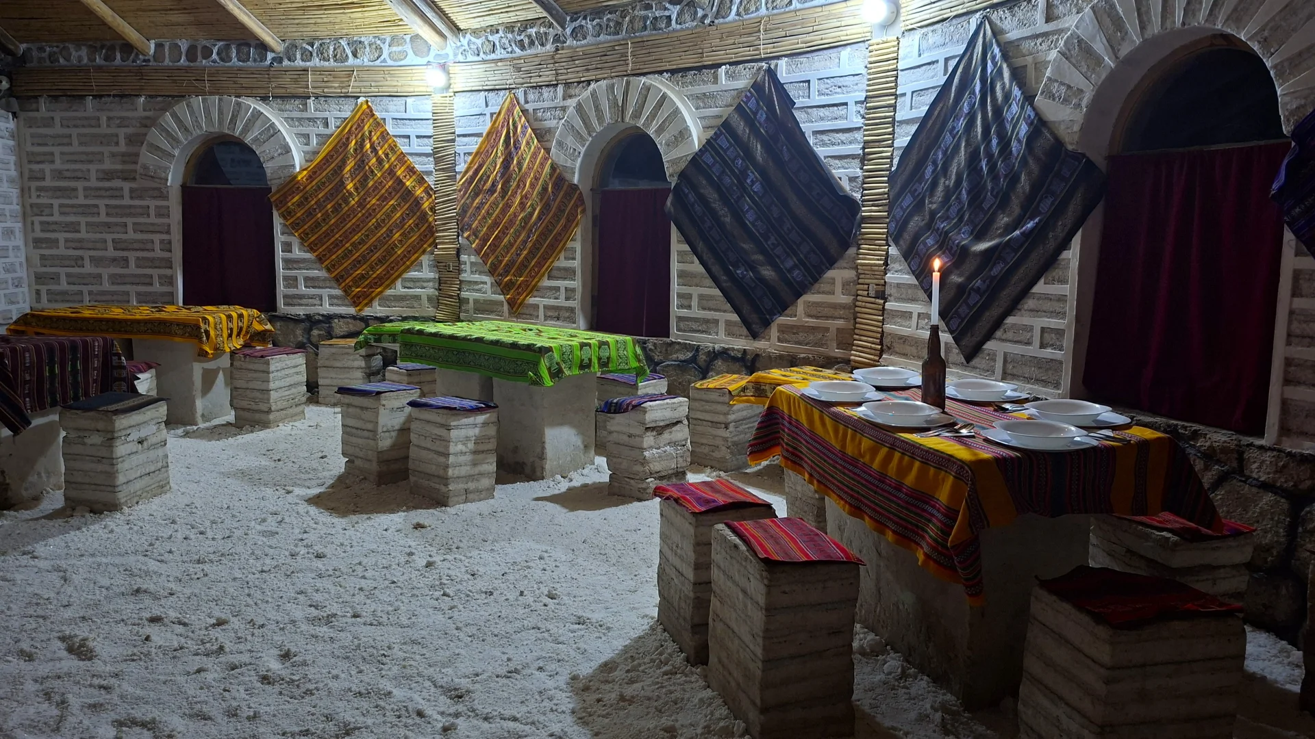 Interior del menjador de l'hotel de sal a San Juan, amb parets, taules i tamborets construïts amb blocs de sal i terra de sal picada, taules decorades amb teixits andins tradicionals de colors vius i una espelma encesa creant un ambient càlid.