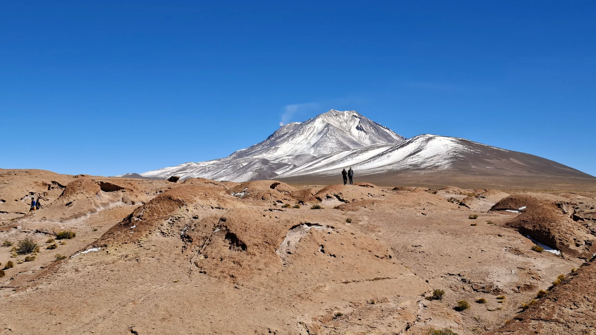 Vista panoràmica del volcà Ollagüe amb els seus cims estriats de neu i una lleugera fumarola blanca que s'eleva des del seu vessant sota un cel blau net, contemplat des d'un turó rocós d'origen volcànic amb petits visitants al cim.